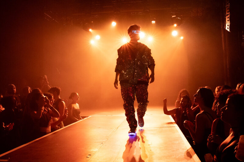 A backlit figure on a stage runway with crowds either wide. The lighting is bright and orange.