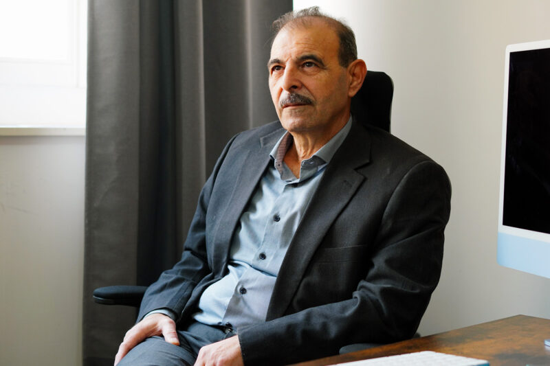 A man in a suit leans back in a chair at his desk, looking contemplatively into the distance. Behind him is a darkened computer monitor