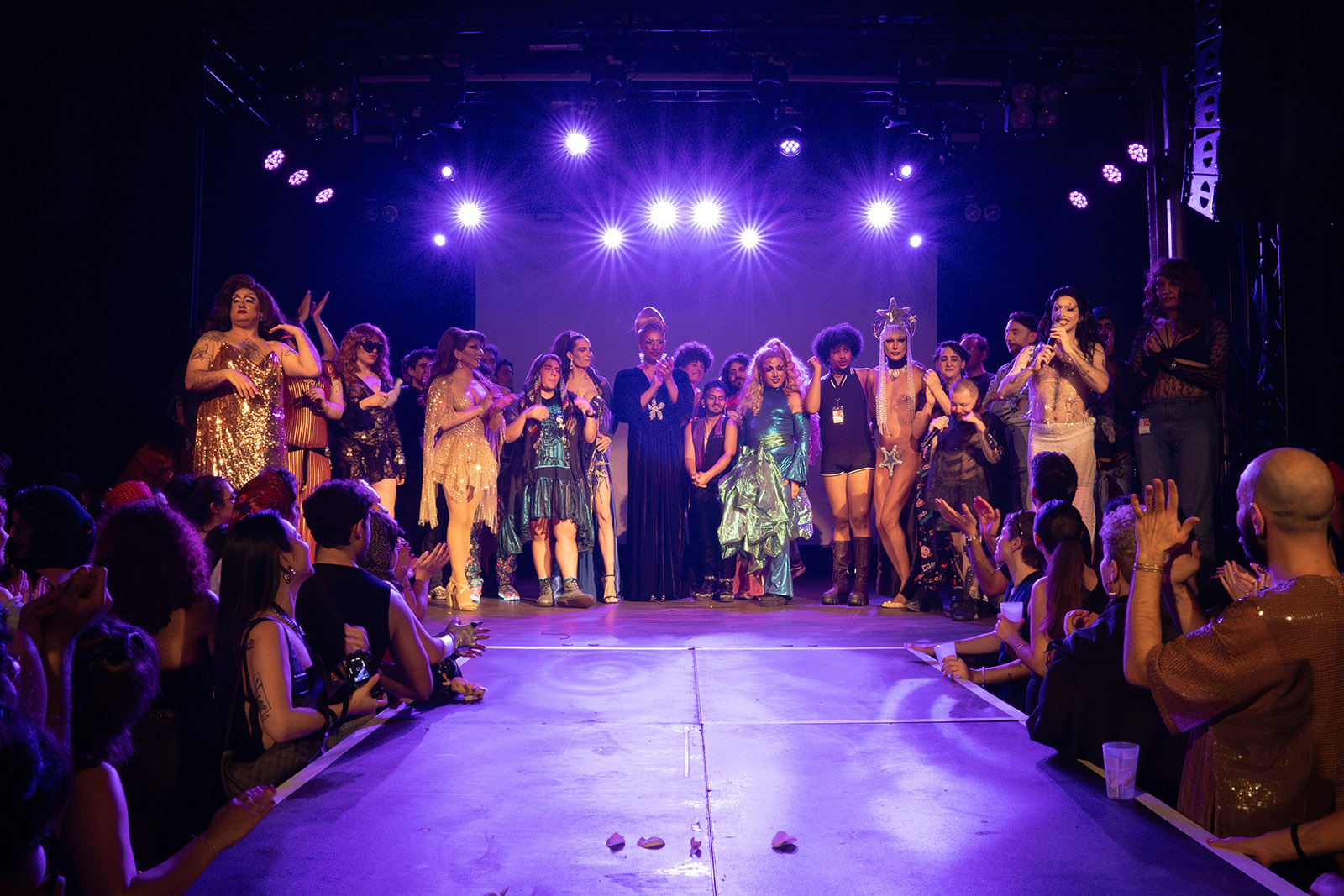 A row of performers on stage, lit by purple lights, many in colourful and shiny costumes. Crowds either side of a walkway look on.
