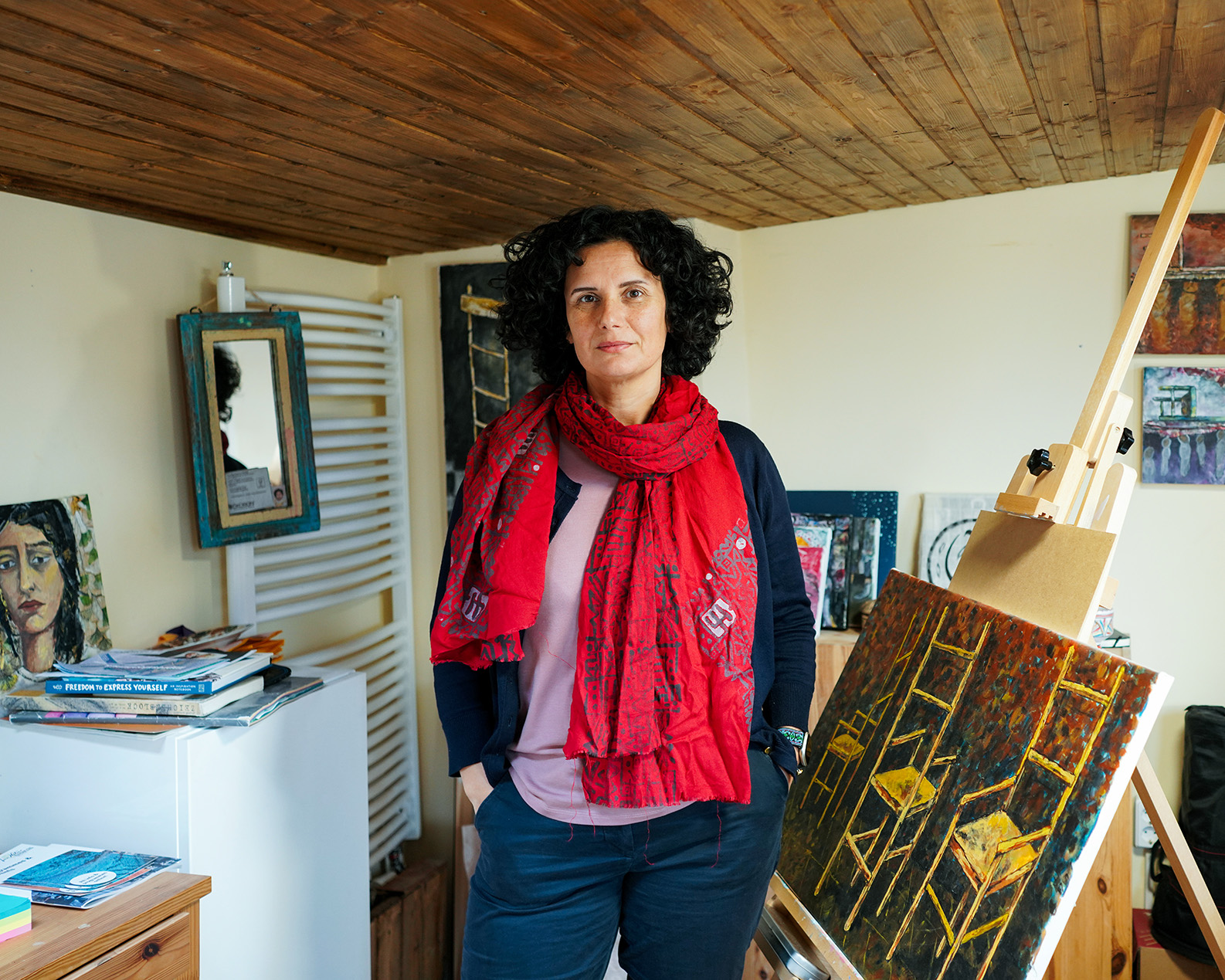 A woman in a red scarf looks neutrally into the camera, surrounded by paintings and books. She is Kefah Ali Deeb, one of a million Syrian refugees in Germany who fled the Assad regime