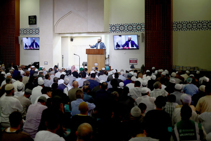 A small suited figure talks and gestures behind a pulpit. A crowd, facing towards him and away from the camera, sits and listens