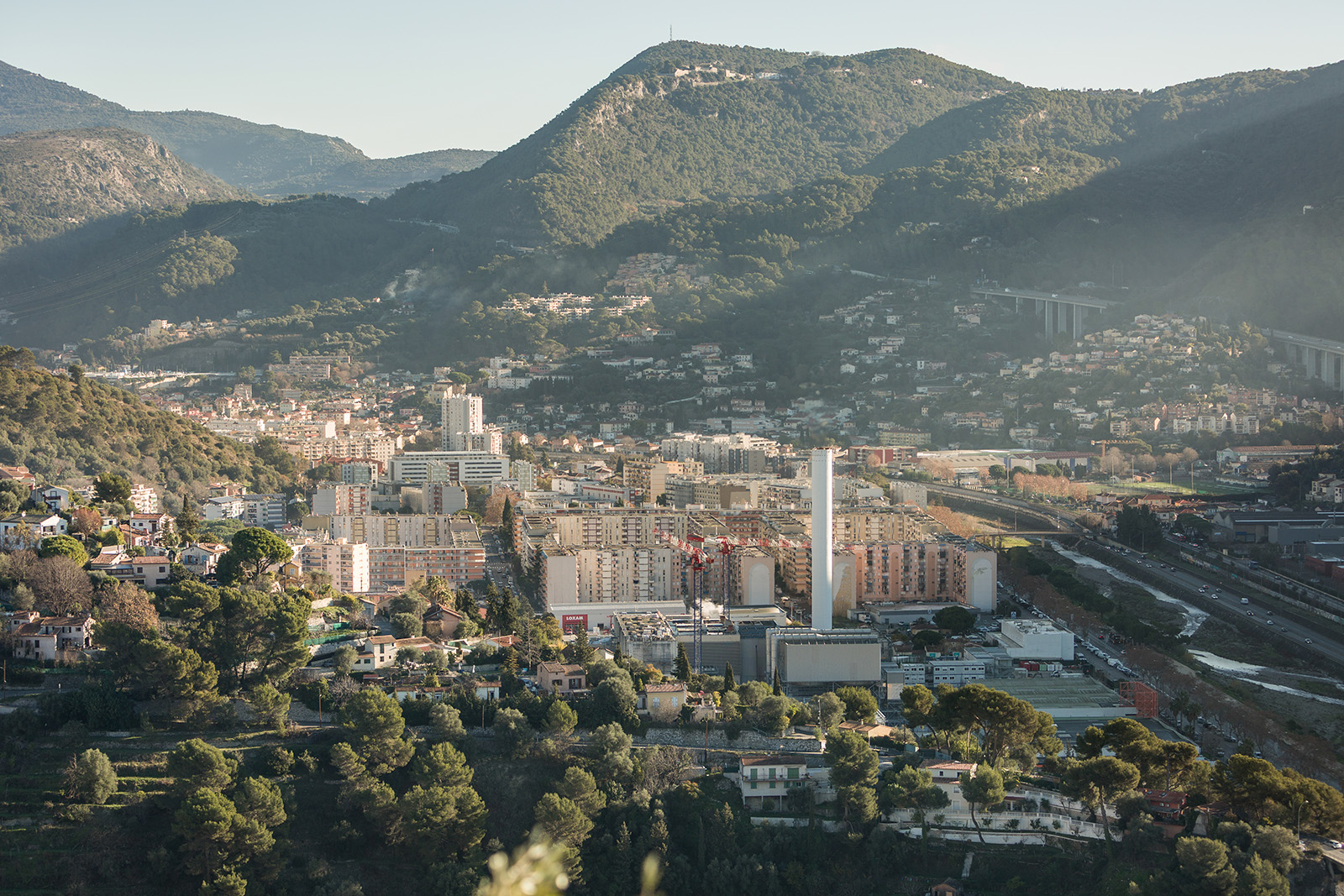A view of a town from a distance, with medium-rise towers arranged at right angles, surrounded by trees and in the distance hills and mountains. The view is sunny
