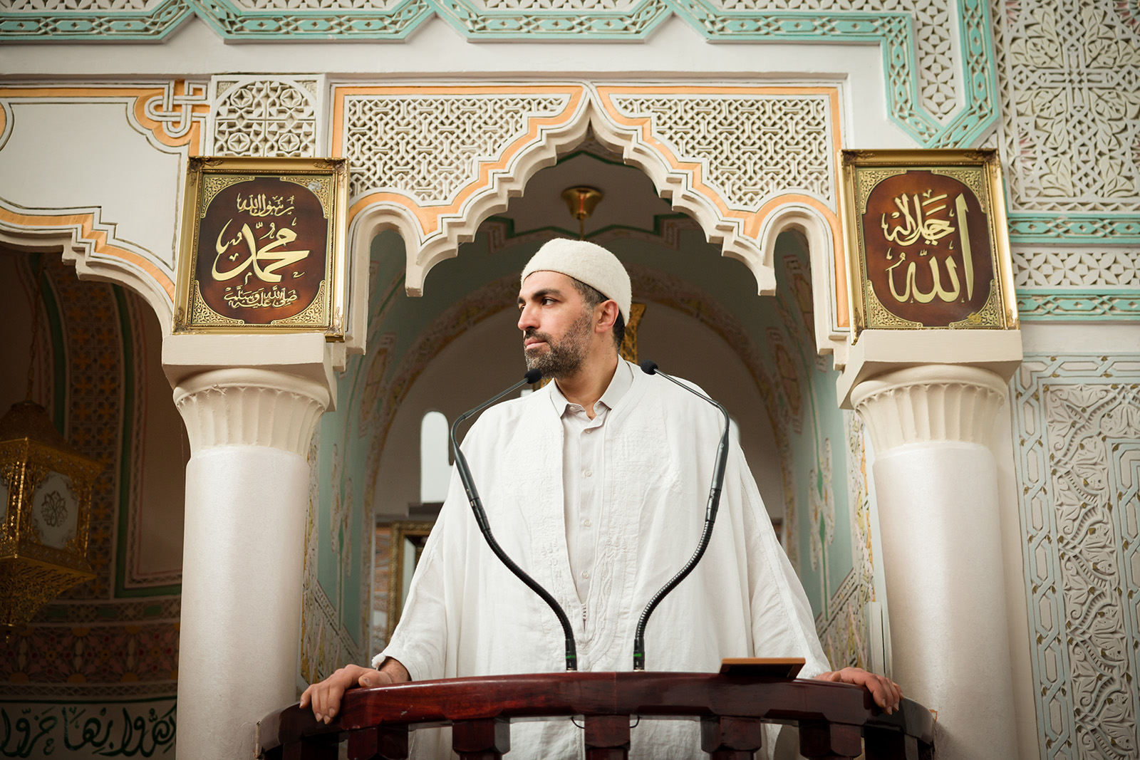 An imam in white looks to one side while gripping the edge of a lectern. His expression is calm. On the walls behind him are Arabic calligraphy and geometric patterns