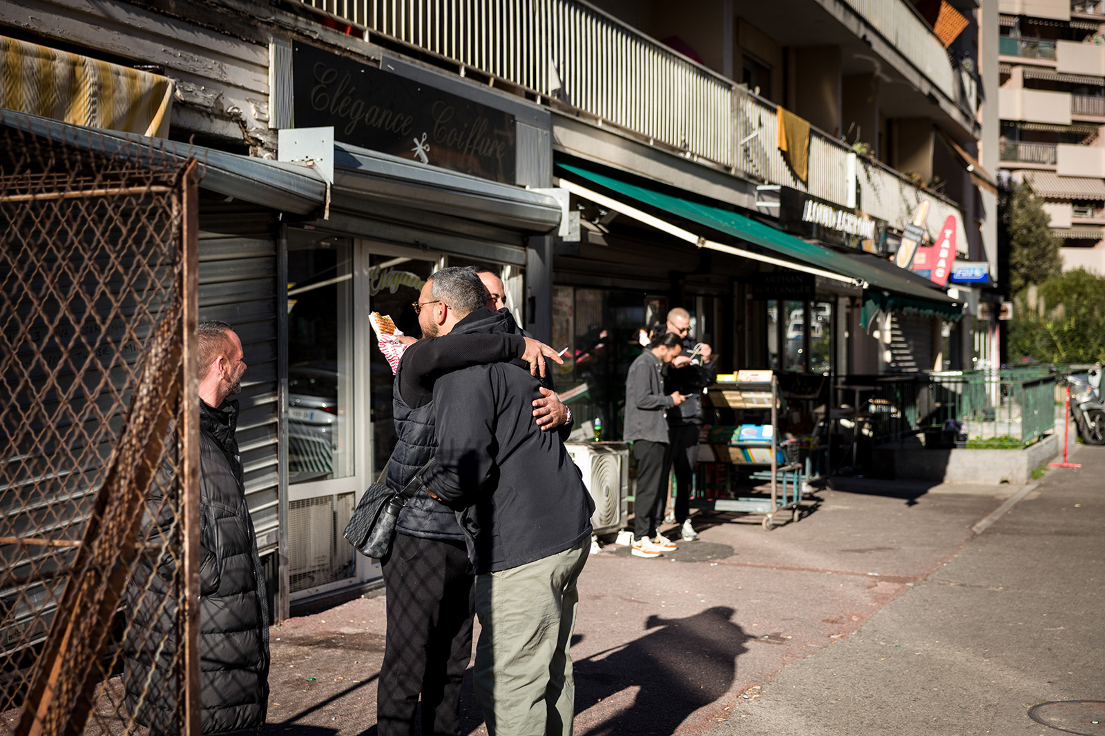 Two people hugging in a sunny street, flanked by shops and grilles