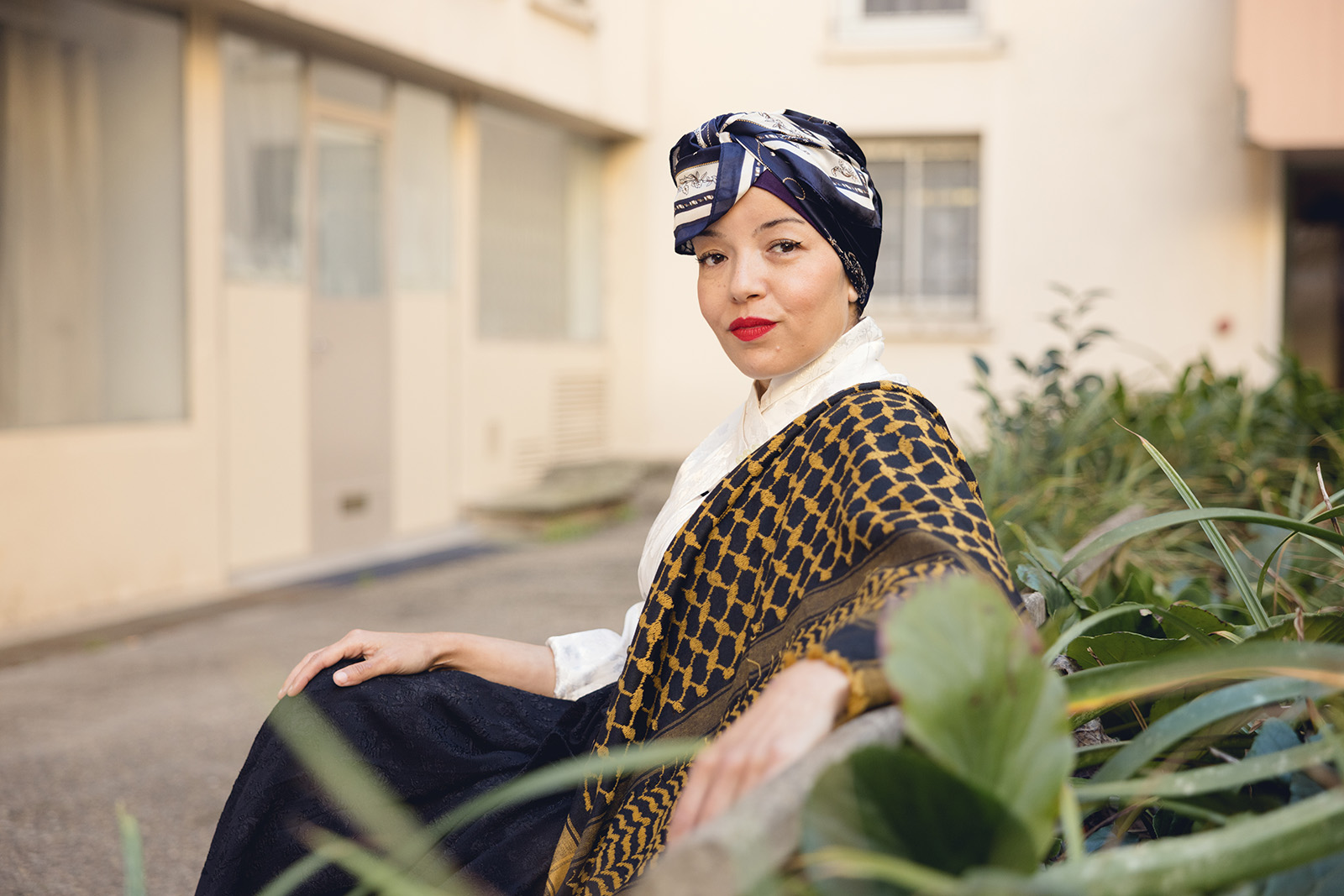 A woman dressed in a patterned top with a silk scarf tied around her hair looks into the camera