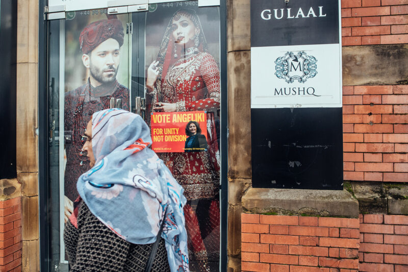 A woman in a hijab walks past the camera while, behind her, a 'vote Labour' poster is visible