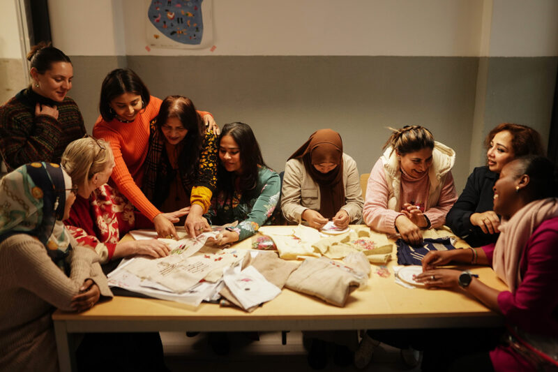 A photograph of a group of women, including Homelore founder Riddhi Varma (4th from left, wearing an orange sweater), gathered round a table laden with embroidery during a workshop
