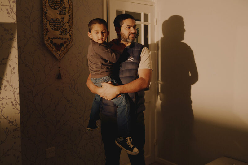 A man holds his young son on his hip. The light is a soft yellowy brown and they are silhouetted against the wall behind.