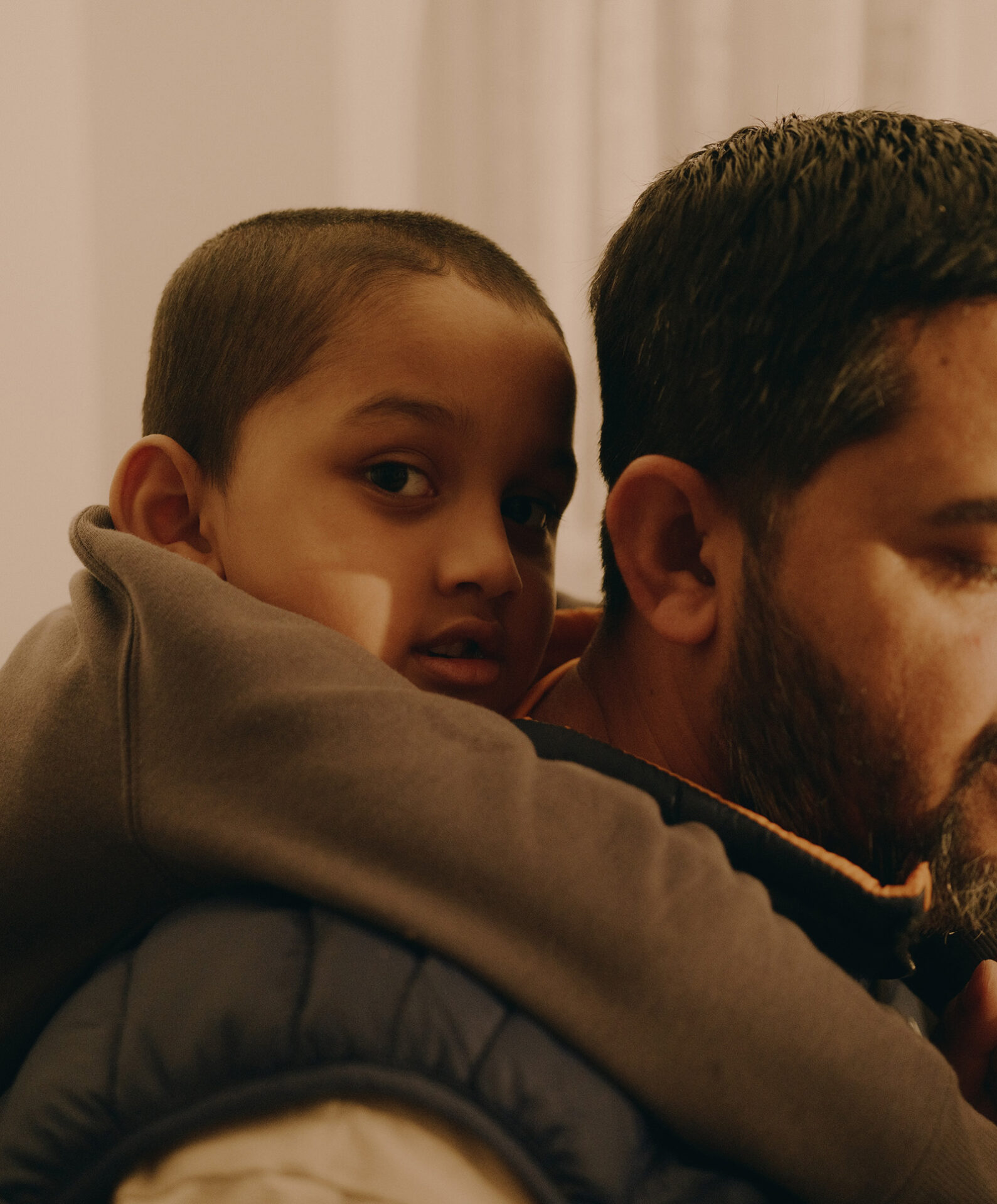 A little boy on his father's shoulders looks into the camera. The father's face is not fully in frame. The light is a warm yellowy brown. The boy's expression is content.