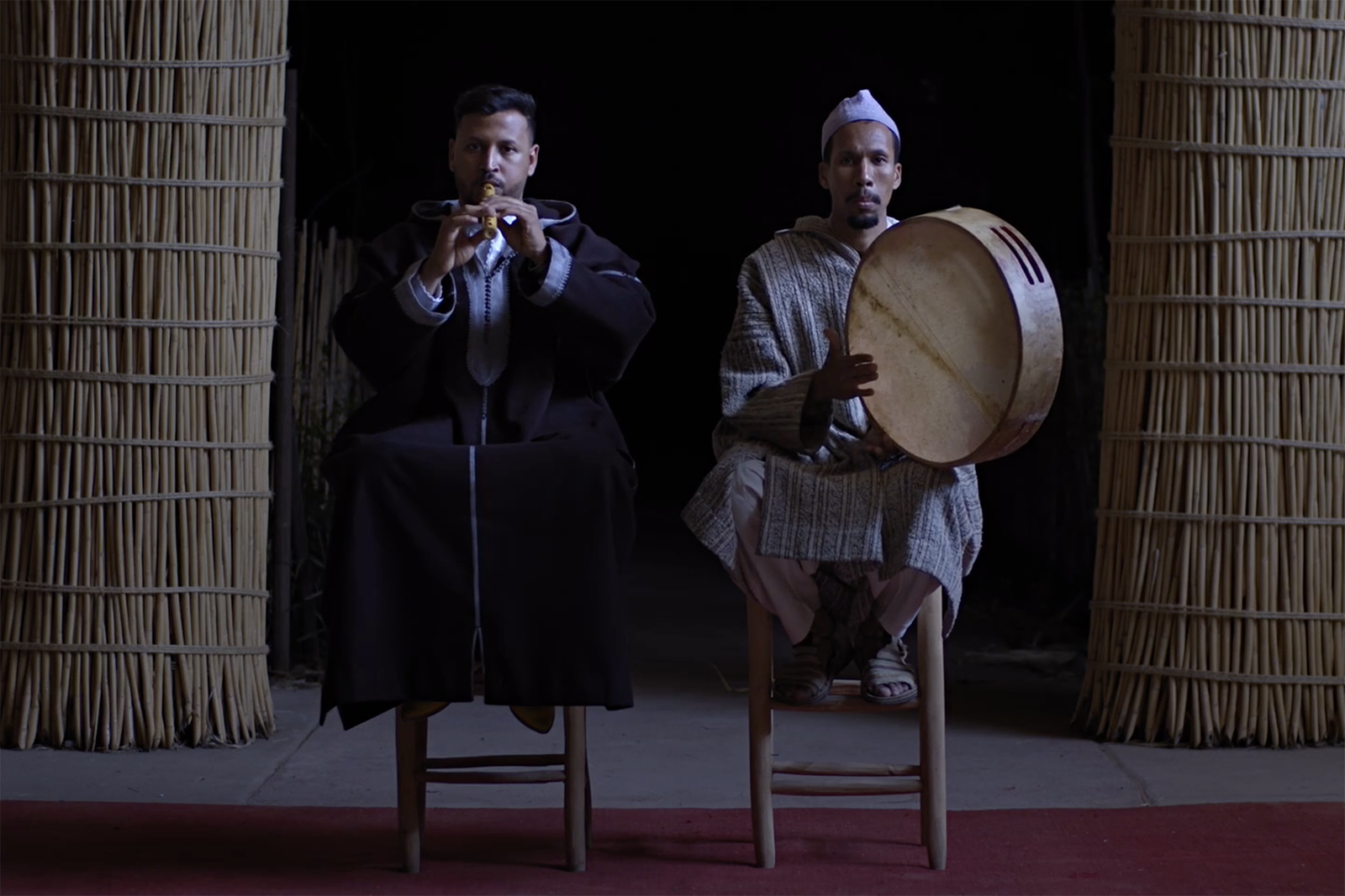 A still image from a video featuring two men sitting side by side on stools, one playing a wooden wind instrument and the other a large tamburine. The video forms part of Bouchra Khalili's Circles and Storytellers, his debut UK solo exhibition