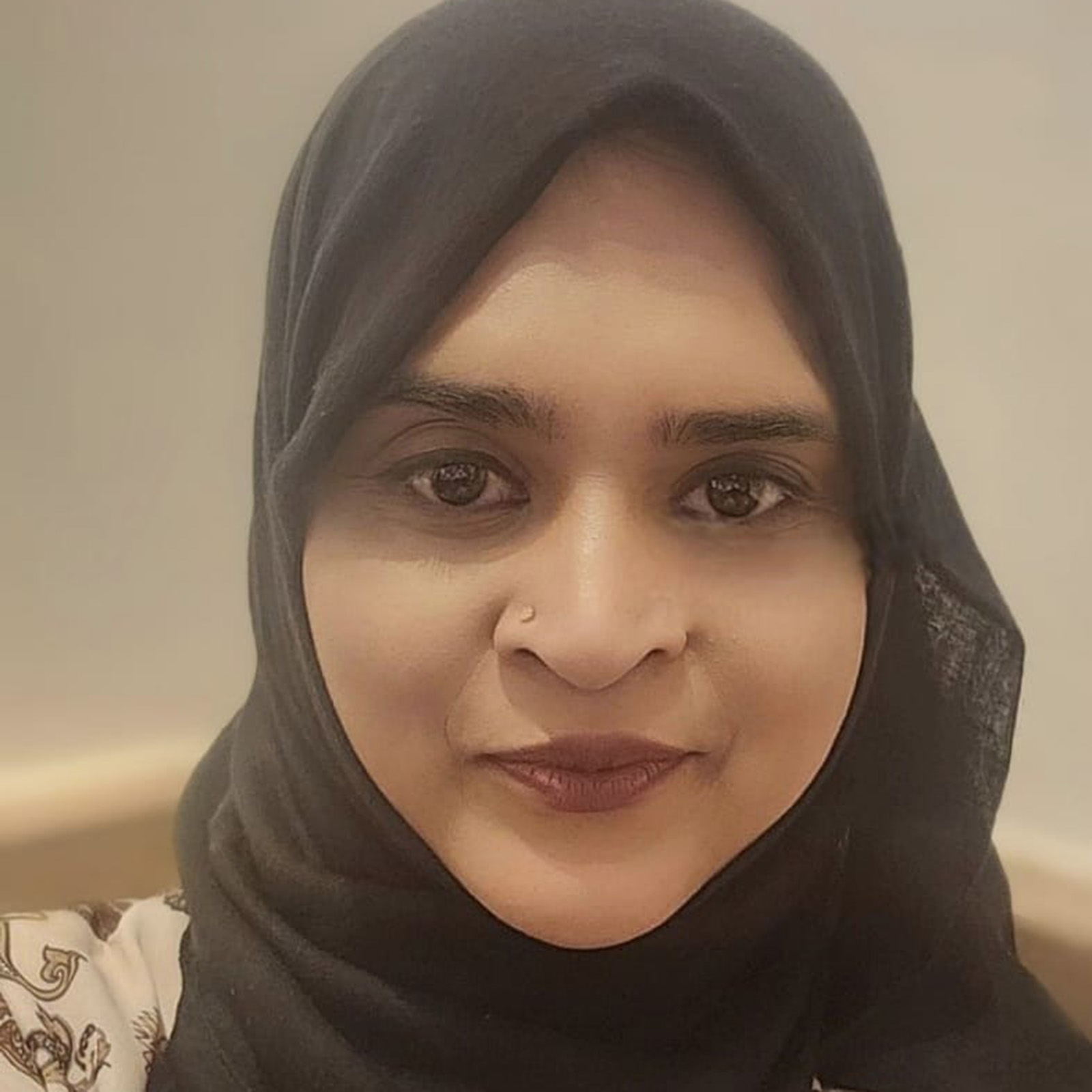 A woman looks directly into the camera with a neutral expression. She wears a black hijab and has a nose piercing.