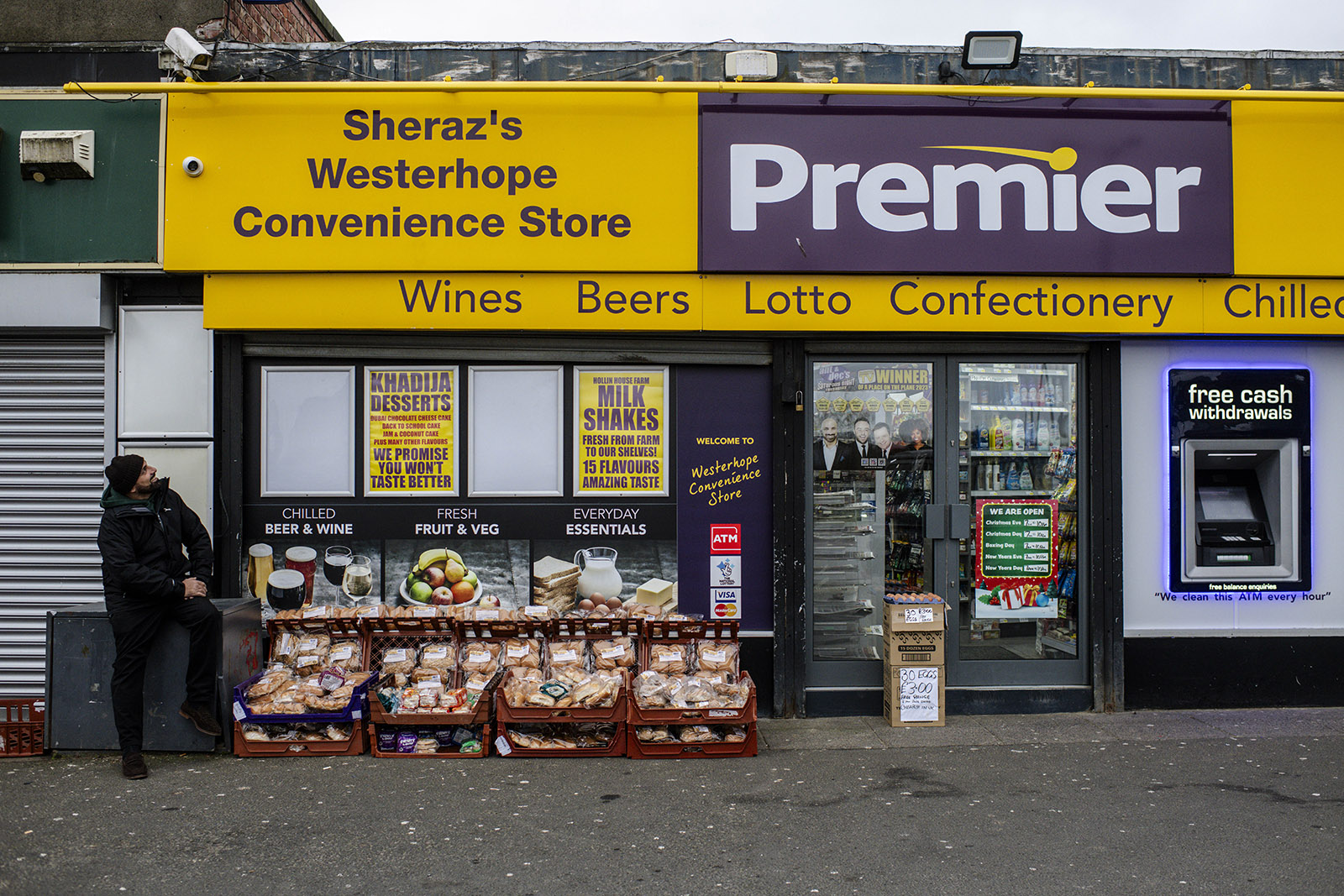 A photograph of Sheraz Awan outside his shop, beside his food giveaway stall, looking up at the store signage and smiling