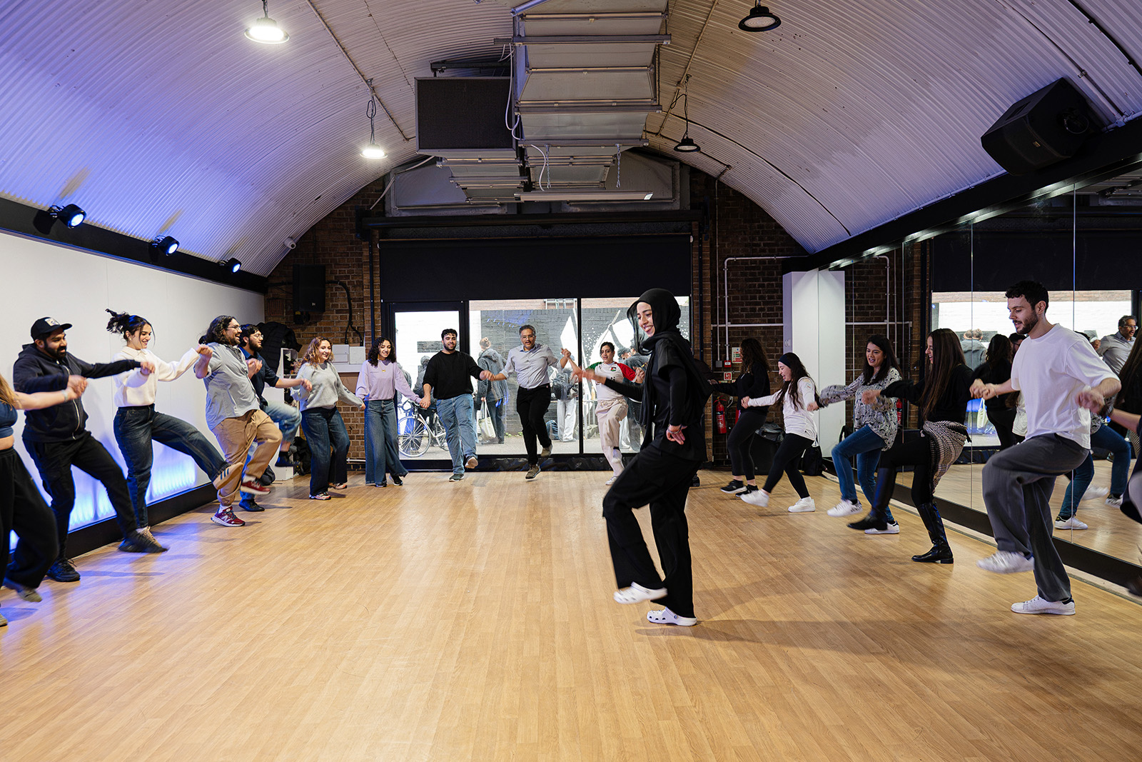 A photograph of a group of dancers linking arms, with an instructor in the middle, at a DabkeRoots workshop taking place in a studio built in an archway in south London