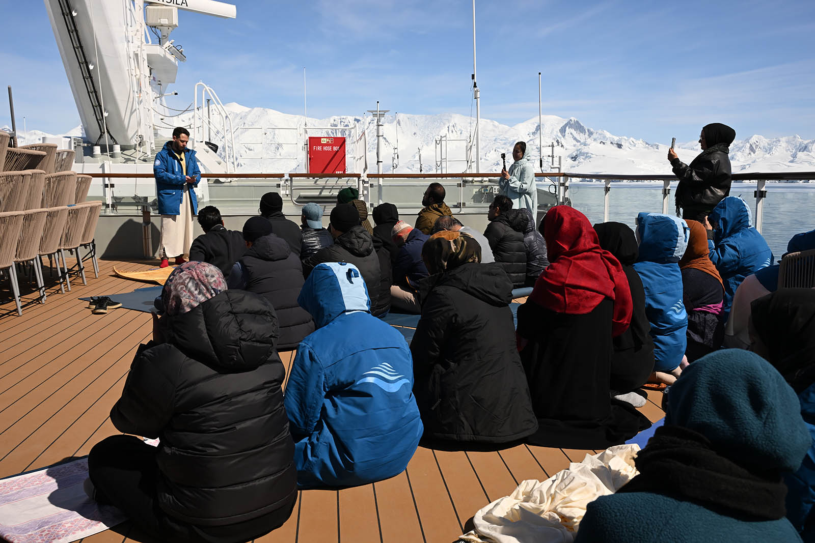 A photograph of Christian performing the khutbah during jummah prayers before a group of seated worshippers on the top deck of the Ocean Albatros, with the coast of Antarctica in the background