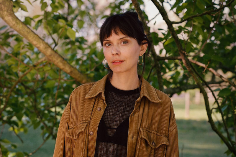 A portrait photograph of author Amber Husain, who is wearing a brown corduroy jacket over a black top and standing in front of a tree