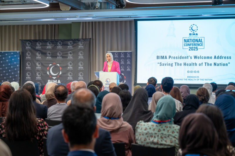 A woman in a pink suit speaks at a lectern to a crowd with their heads facing her, away from the camera. A screen beside her reads 'Bima president's welcome address: saving the health of the nation'