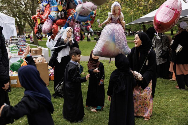 A photograph showing a group of women and children, some holding brightly coloured balloons — one in the shape of a fairytale princess — celebrating Eid al-Fitr during a 1Eid event in Southall Park, London in April 2024