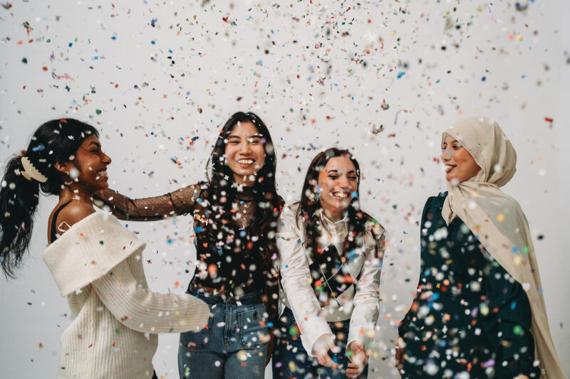 A stock photograph of a diverse group of young women cheerfully smiling, laughing and enjoying a celebration party with colourful falling confetti on a white background