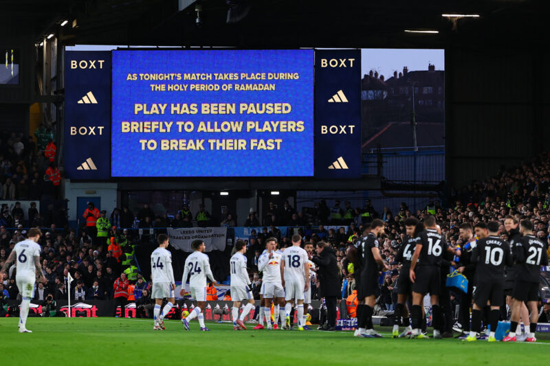 A photograph of a message displayed on a large LED screen in Leeds United's Elland Road stadium on Saturday, 28 February 2026, stating: 'Tonight's match takes place during the holy period of Ramadan. Play has been paused briefly to allow players to break their fast." In the foreground, on the pitch, players from the home team and their opponents Manchester City gather on the touchline for a drinks break.