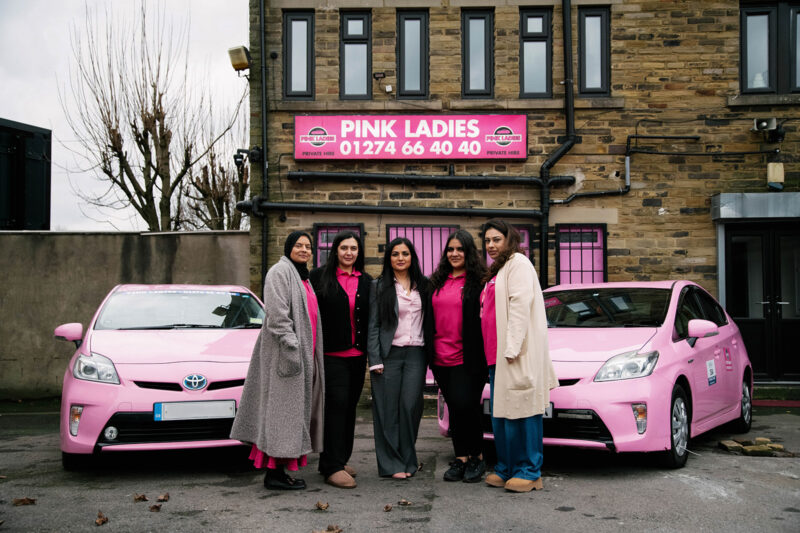 Photograph of five members of the team of the cab company. Pink Ladies sign in the background with two pink cars. Amberine Nawaz (centre) with some of the Pink Ladies team outside their office. In the office the drivers checked in with each other about different riders or journeys.