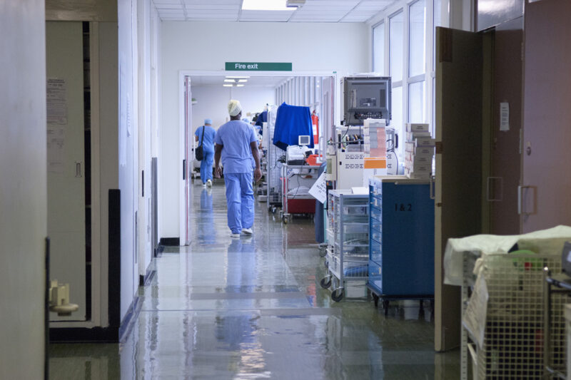A stock image of a hospital corridor, with two health workers in sanitary clothing walking away from the camera