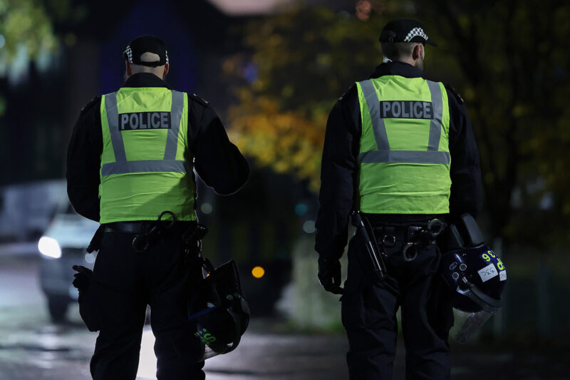 Two police officers with their backs turned. One holds a helmet.