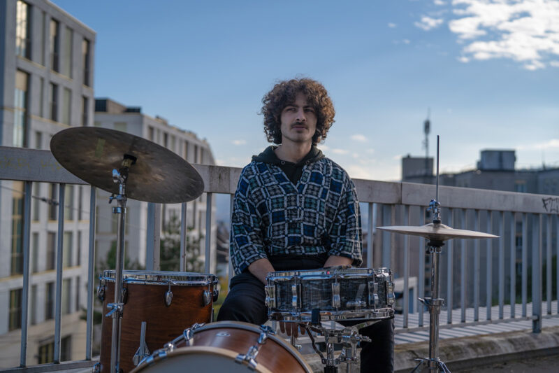 A photograph of Swiss-Palestinian drummer and bandleader Ramzi Hammad, seated behind a drum kit on a building roof terrace