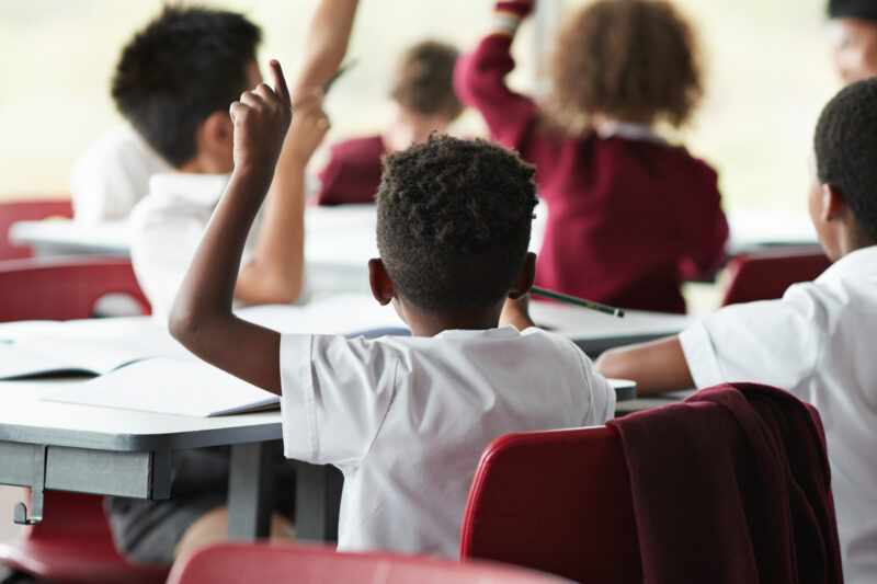 Young children facing away from the camera sit at school desks with their hands raised