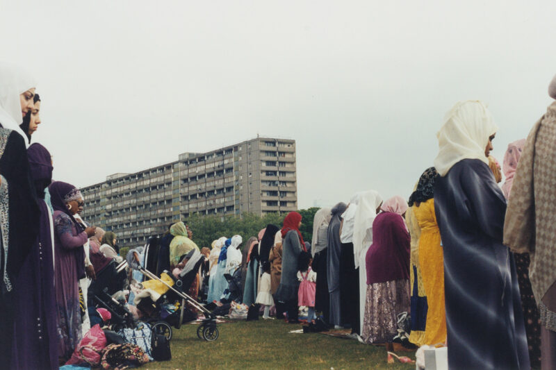 A photograph of women and children standing to perform Eid prayer in Burgess Park, south London, with a block of flats in the background. The image forms part of photographer Zaineb Abelque's project Inside, Outside.