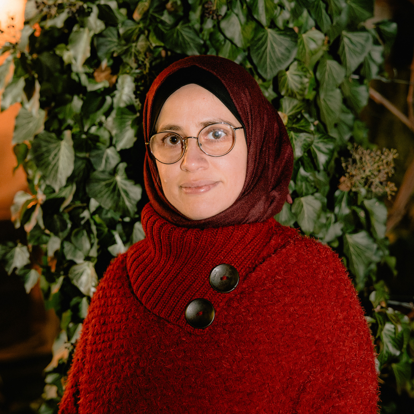 A portrait photograph of Hadil Hafar, wearing a red jumper and headscarf, outside of the family home on the outskirts of Vienna, Austria.
