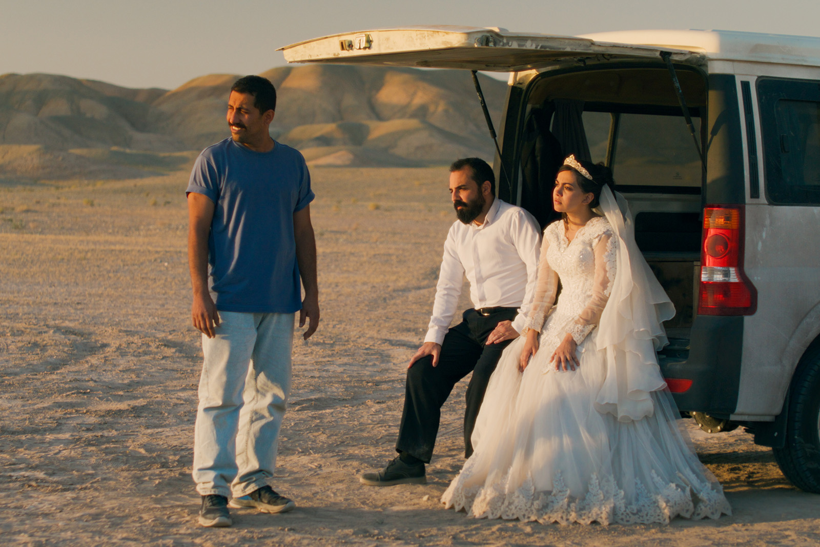 A still image from Jafar Panahi's It Was Just an Accident, which is up for best international feature at the 2026 Oscars. The image features two men and a woman in a wedding dress, by a vehicle in the desert.