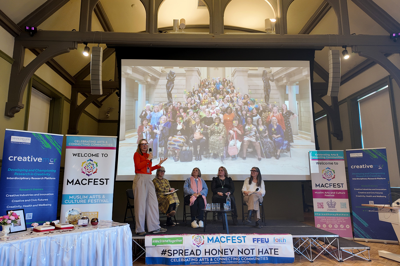 A photograph of a panel of five women on a low stage, one on the left standing to speak and the others seated, in front of a big screen showing a still image of a large, diverse group of women. The occasion is a MACFEST IWD 2026 event at the Whitworth art gallery in Manchester on 4 March 2026
