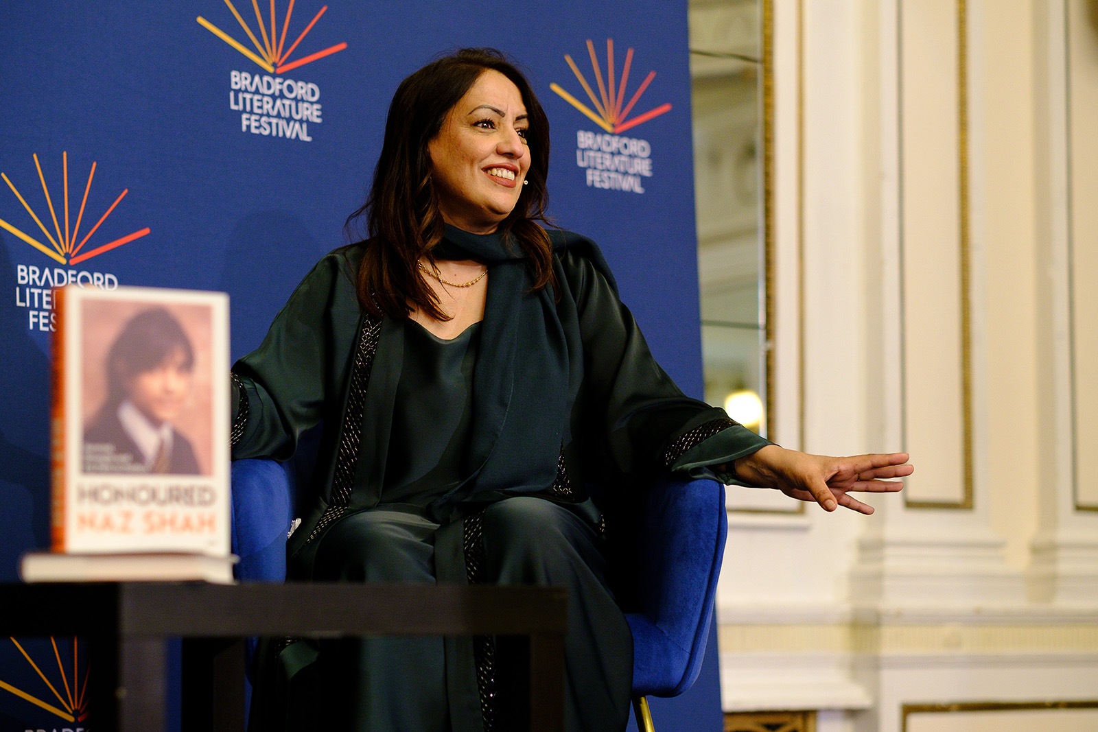 Naz Shah, pictured seated on a stage with a copy of her book Honoured propped up on a table in front of her, at the title's launch event at the Bradford Literature Festival 2026