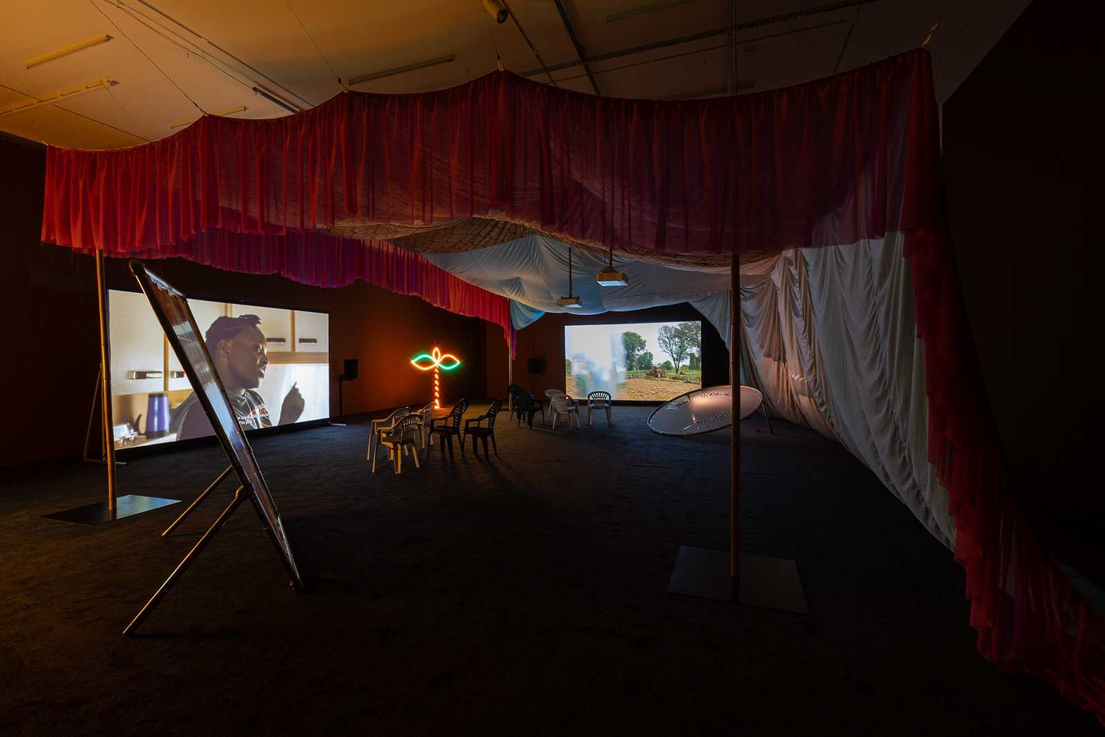 A photograph of Rehana Zaman's Plantation exhibition at Sheffield's Site Gallery, showing a viewing area enclosed by a large drape suspended from the ceiling, with chairs and two big screens inside