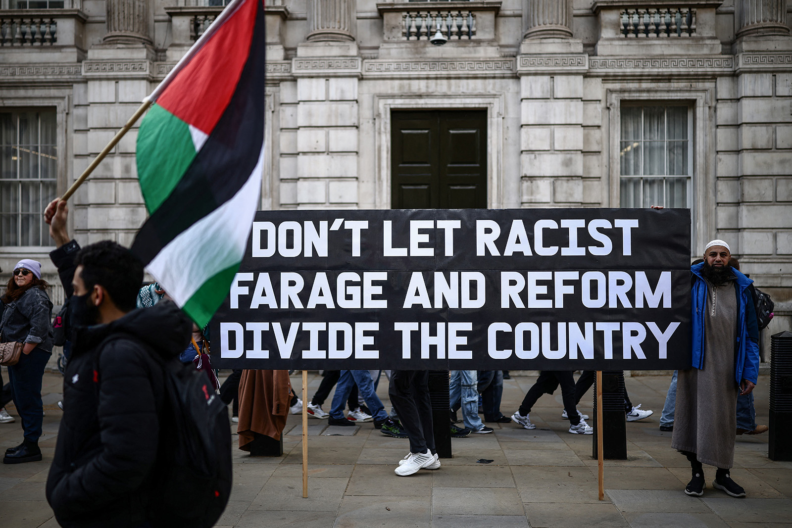 A man waves a Palestinian flag in front of protesters displaying a placard, 'Don't Let Racist Farage and Reform divide the country', during the Together Alliance rally in central London on 28 March 2026.
