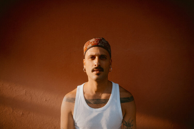 A portrait photograph of Palestinian singer-songwriter Ahmed Eid, wearing a white vest that shows his tattooed shoulders and looking directly at the camer