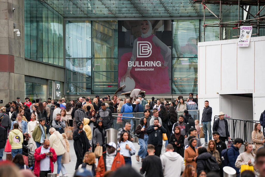 A crowd mills about in front of the entrance to a shopping centre. Above them are glass panels with a banner in the middle reading 'Bullring Birmingham'