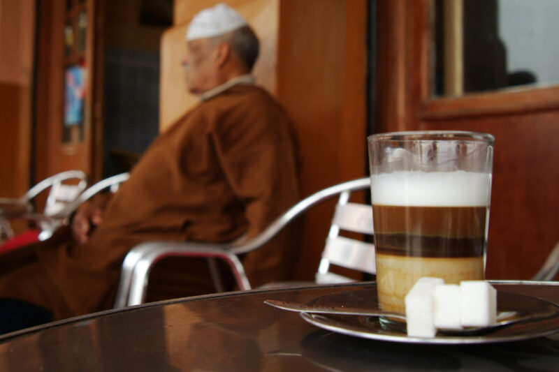 A close-up image of a glass of qahwa nus-nus, served with lumps of sugar and a teaspoon on a saucer, in a Marrakech cafe