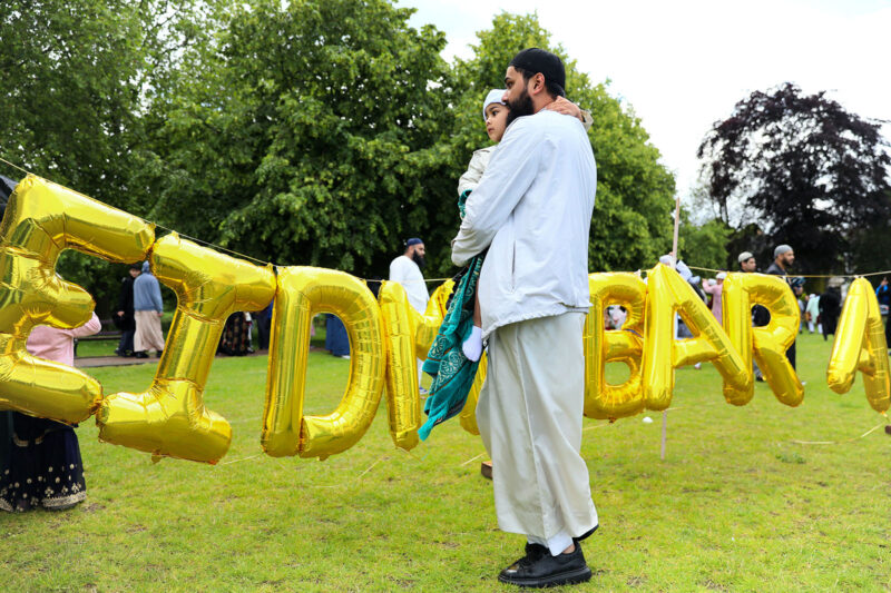A man holds a child in a park. Behind them are gold-coloured balloons reading EID MUBARAK