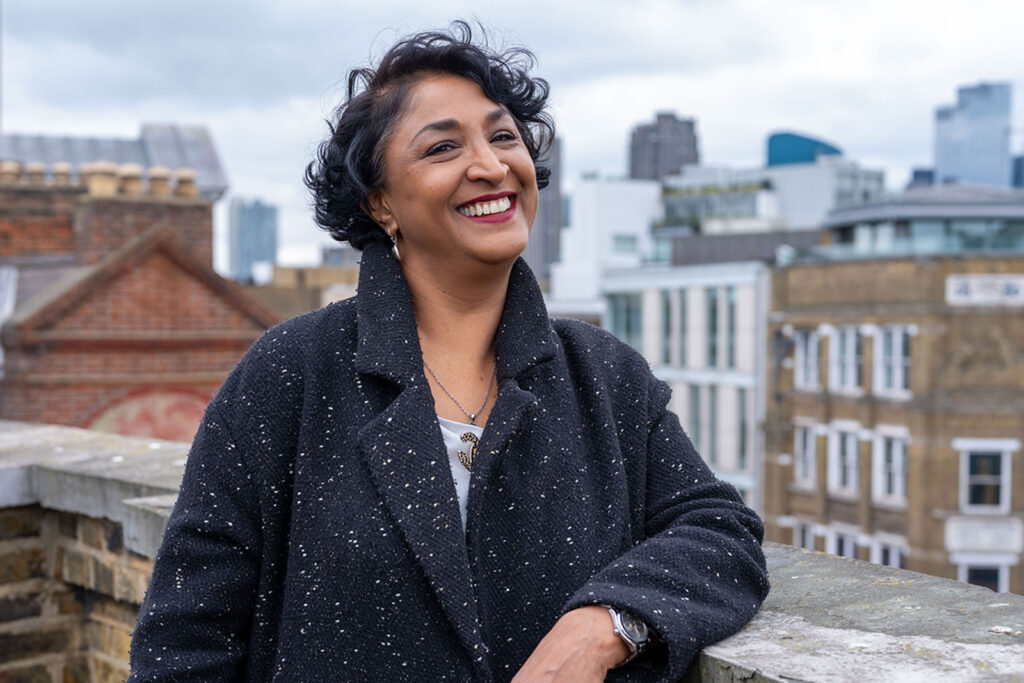 A photograph of fashion tailor Nafisa Tosh, leaning against a rooftop parapet in central London and smiling at the camera