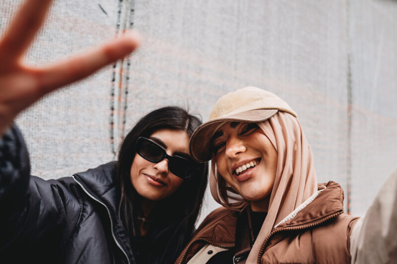 A stock photograph of two young adult girls posing for the camera, one wearing a fashionable hijab with a hat.