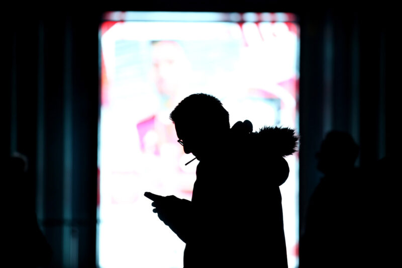 A silhouette of a man against an out-of-focus ad display, smoking and looking at his phone