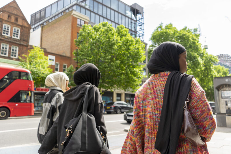 A stock image of a young women wearing hijabs walking down a street, with a red Boris bus in the background