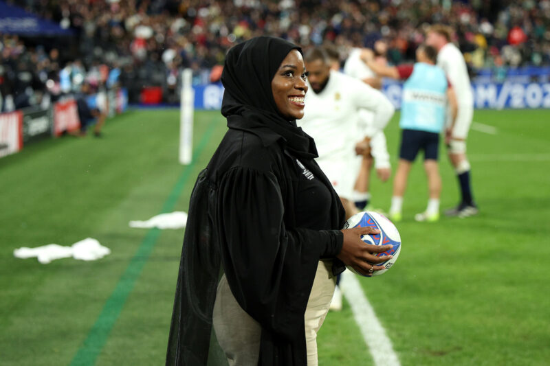 A photograph of a smiling Zainab Alema, waiting pitchside to present the match ball to the referee, ahead of the England v South Africa men's Rugby World Cup semi final in Paris on 21 October 2023