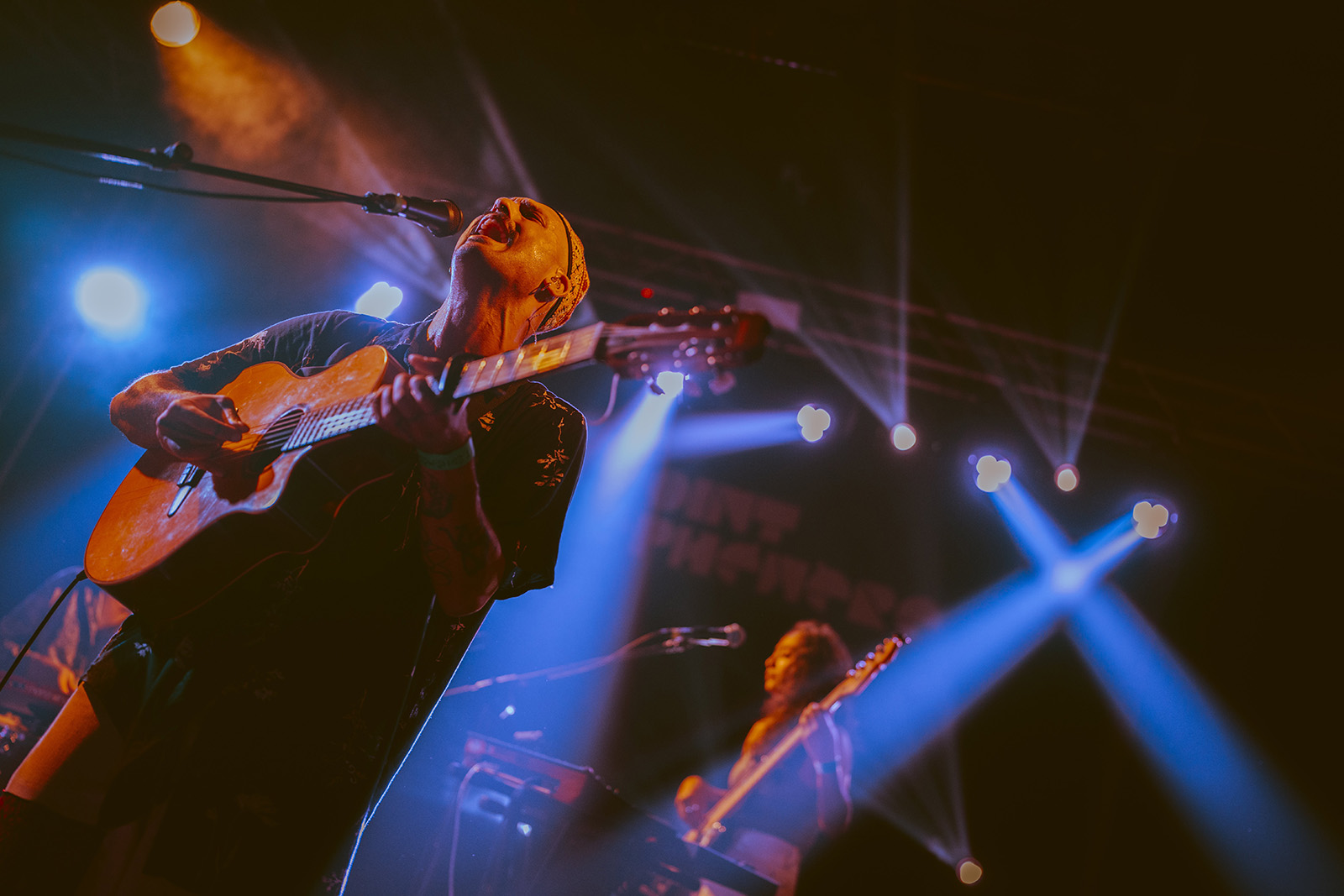 A photograph of Ahmed Eid performing at a gig, singing into a mic while playing an acoustic guitar, taken in 2024