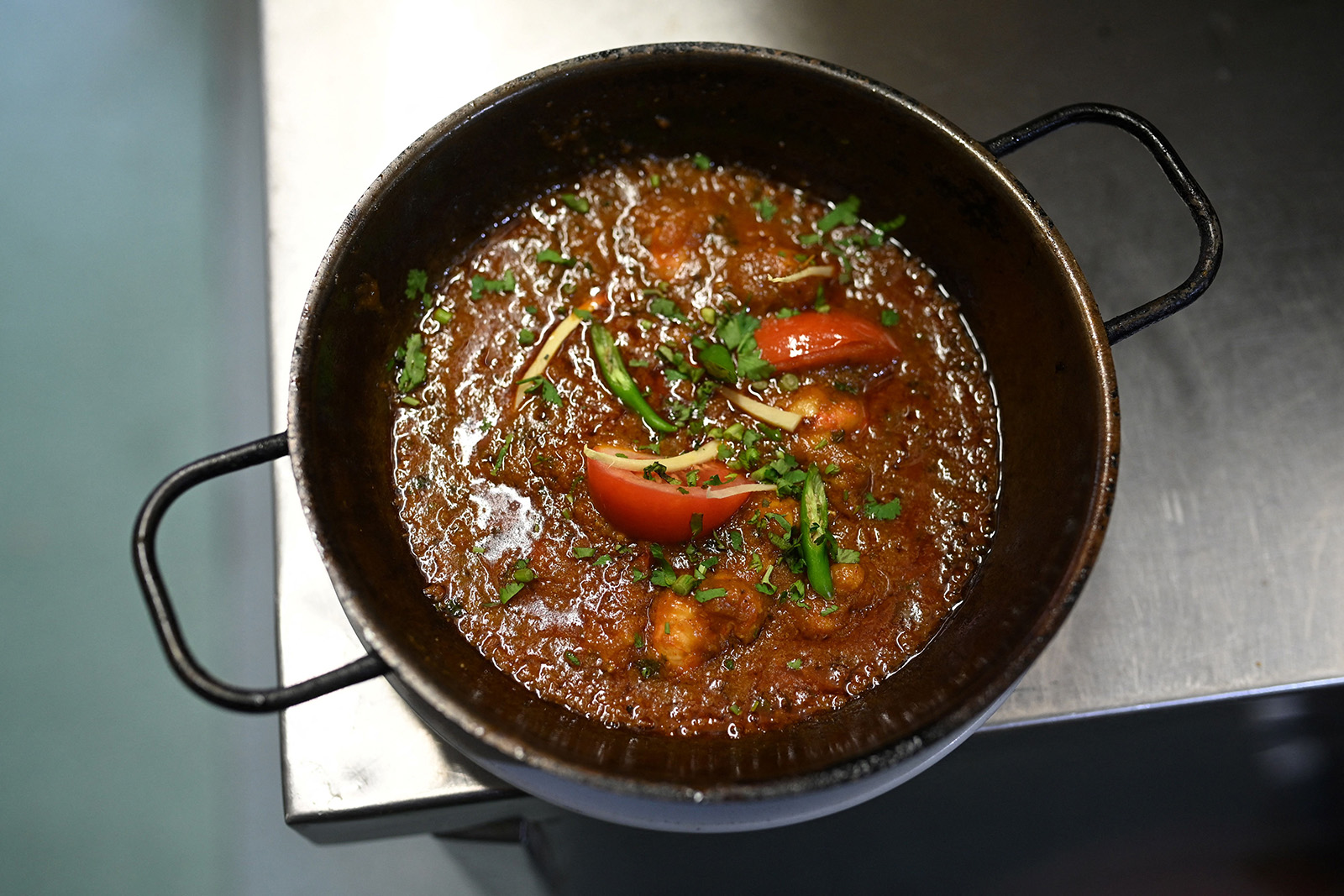 An overhead photograph of balti dish by Birmingham restaurant Shahi Nan, with green chillis and coriander sprinkled over king prawns and tomatoes in a sauce, presented in a two-handled wok