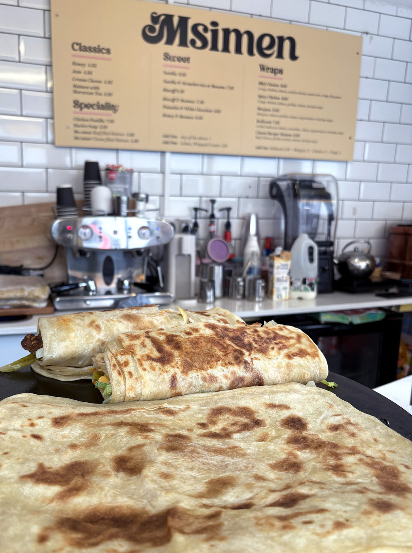 A photograph taken inside Msimen, west London. Msemen, traditional Moroccan pancakes, are pictured in front of a food preparation counter and a menu board on the wall