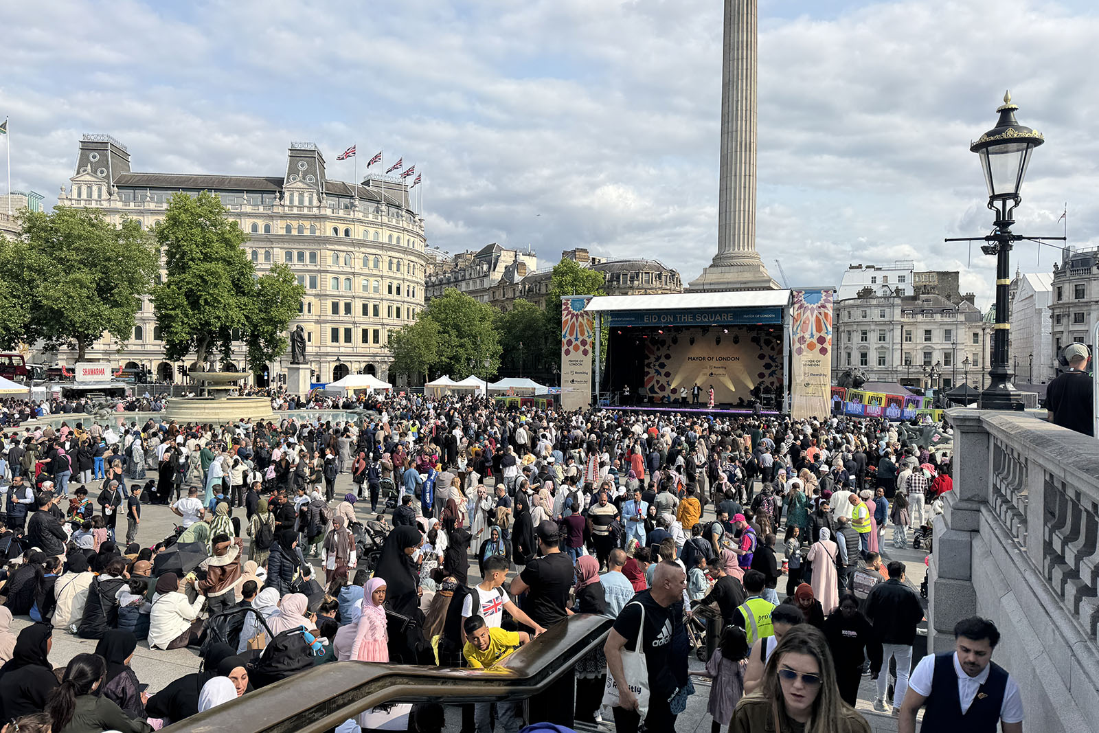 A crowd in the sun, sitting and standing, with Nelson's Column visible behind a temporary stage.