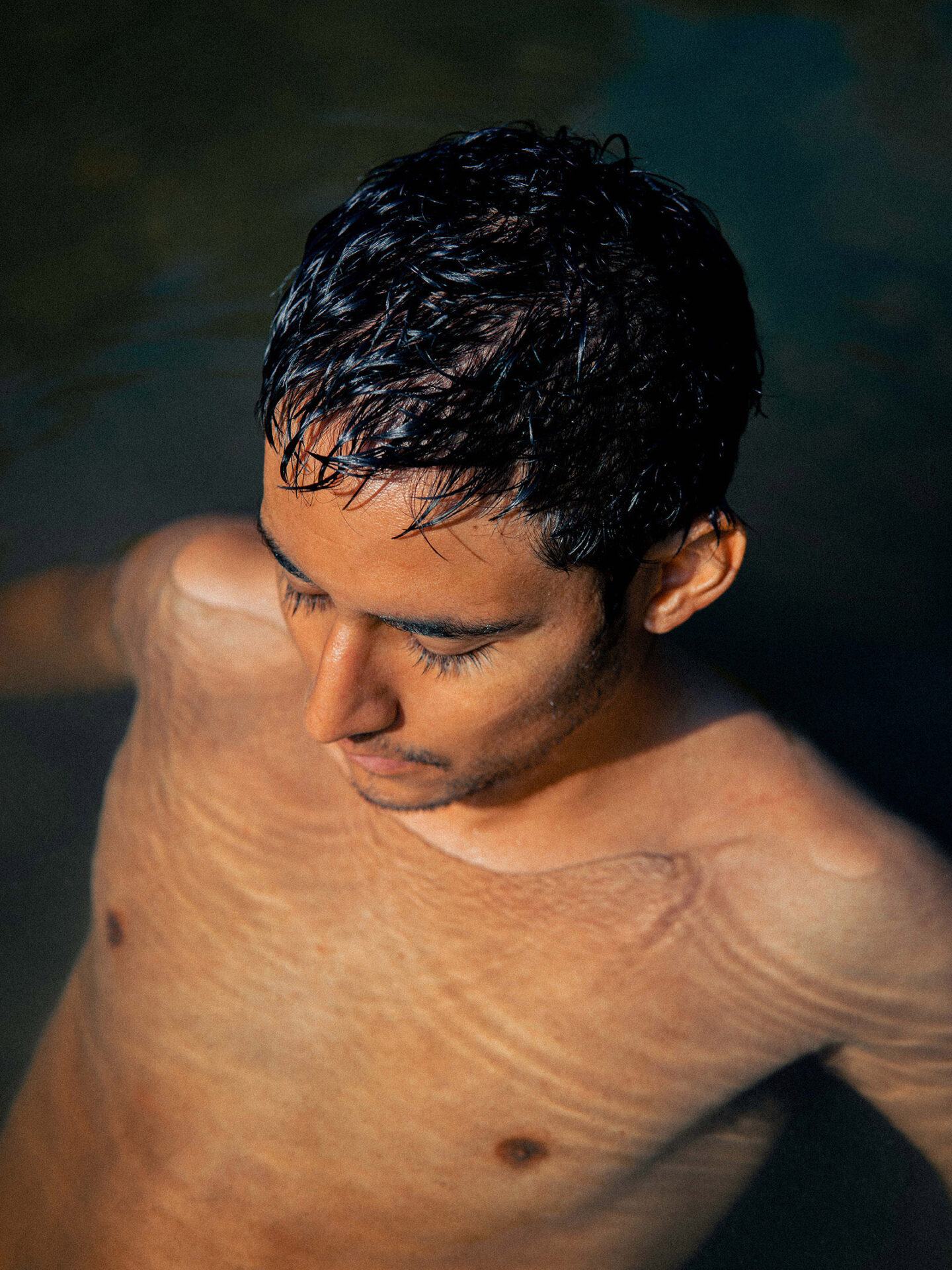 An image taken from above of a young man submerged in water up to his neck, bathed in sunlight