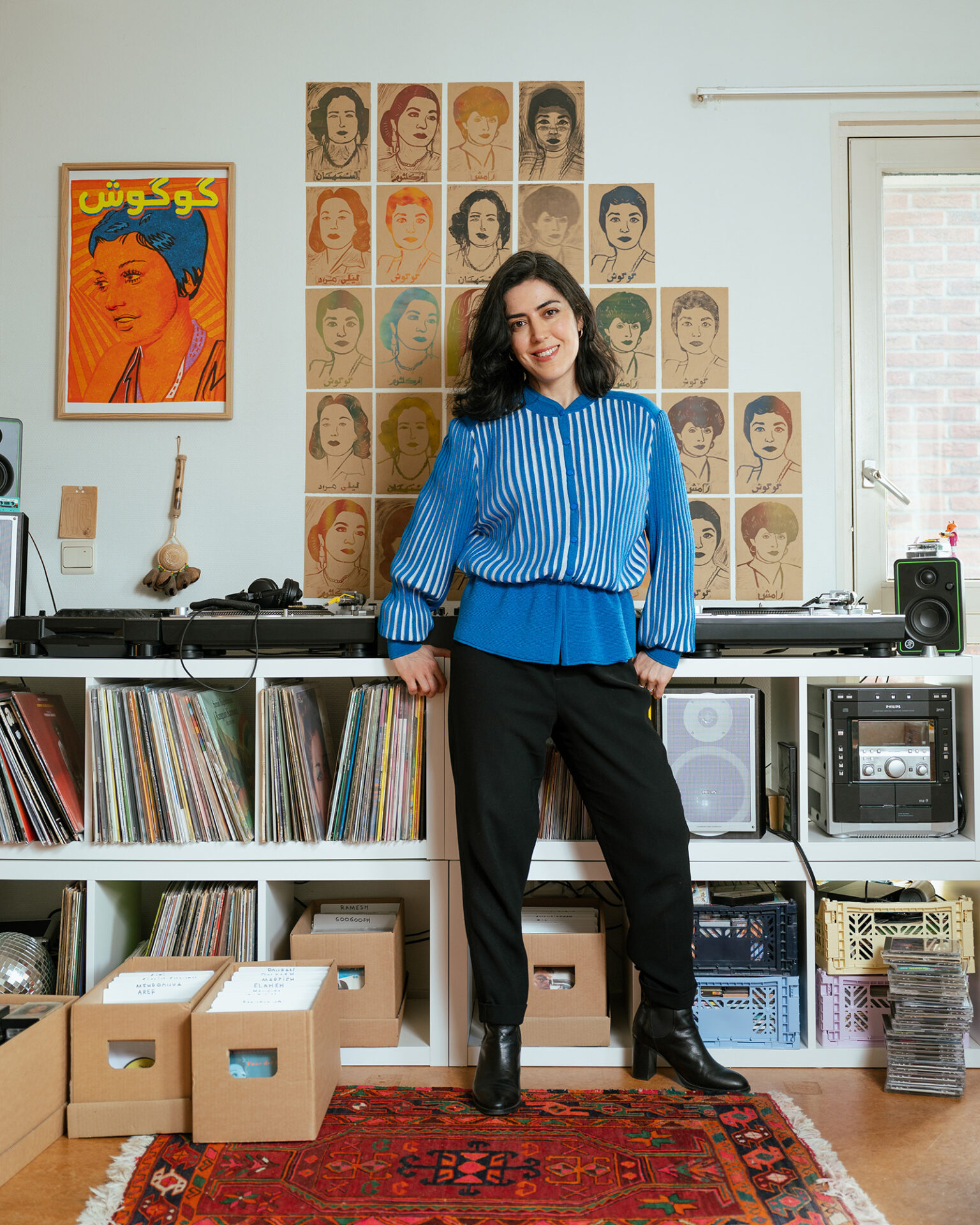 A portrait photograph of Katayoun Arian, who DJs as Katayoun and runs the Disco Diaspora night in Amsterdam, at home standing in front of her record collection and turntables